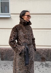 Manteau en fausse fourrure imprimé léopard avec col montant, poches latérales et ceinture à nouer en cuir noir. Texture douce et tons de couleur chauds.