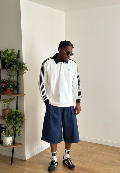 Hombre con gafas de sol, camiseta blanca de manga larga Adidas con rayas negras, pantalones cortos anchos vaqueros, calcetines a rayas y zapatillas negras, de pie en el interior cerca de plantas.