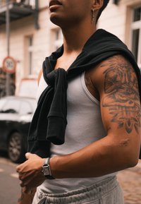 Μαύρος πουλόβερ δεμένος γύρω από τον λαιμό, λευκό μπλουζάκι, γκρι φόρμες, ασημένιο ρολόι και ένα λεπτομερές τατουάζ στον βραχίονα. Αστική σκηνή στο παρασκήνιο.