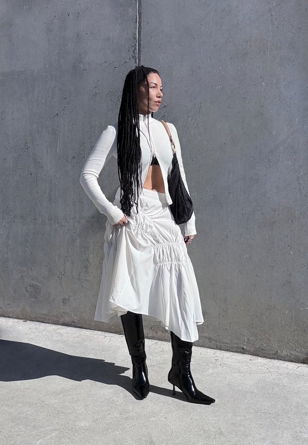 Femme aux longues tresses, debout contre un mur en béton, portant un haut court blanc, une jupe blanche froncée, des bottes noires et un sac à bandoulière noir.