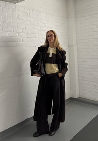 Čierny kabát cez béžový a hnedý pruhovaný sveter, široké čierne nohavice a čierne mokasíny. Modelka drží čiernu kabelku a má okuliare.