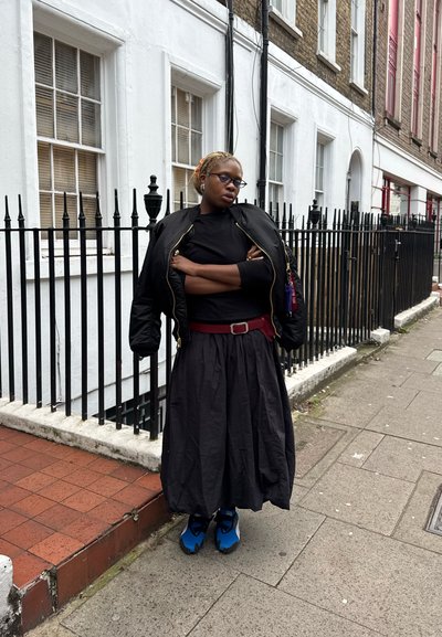 Chaqueta bomber negra, top negro y falda larga negra; zapatos azules con detalles en negro. De pie frente a un edificio blanco y una valla metálica.