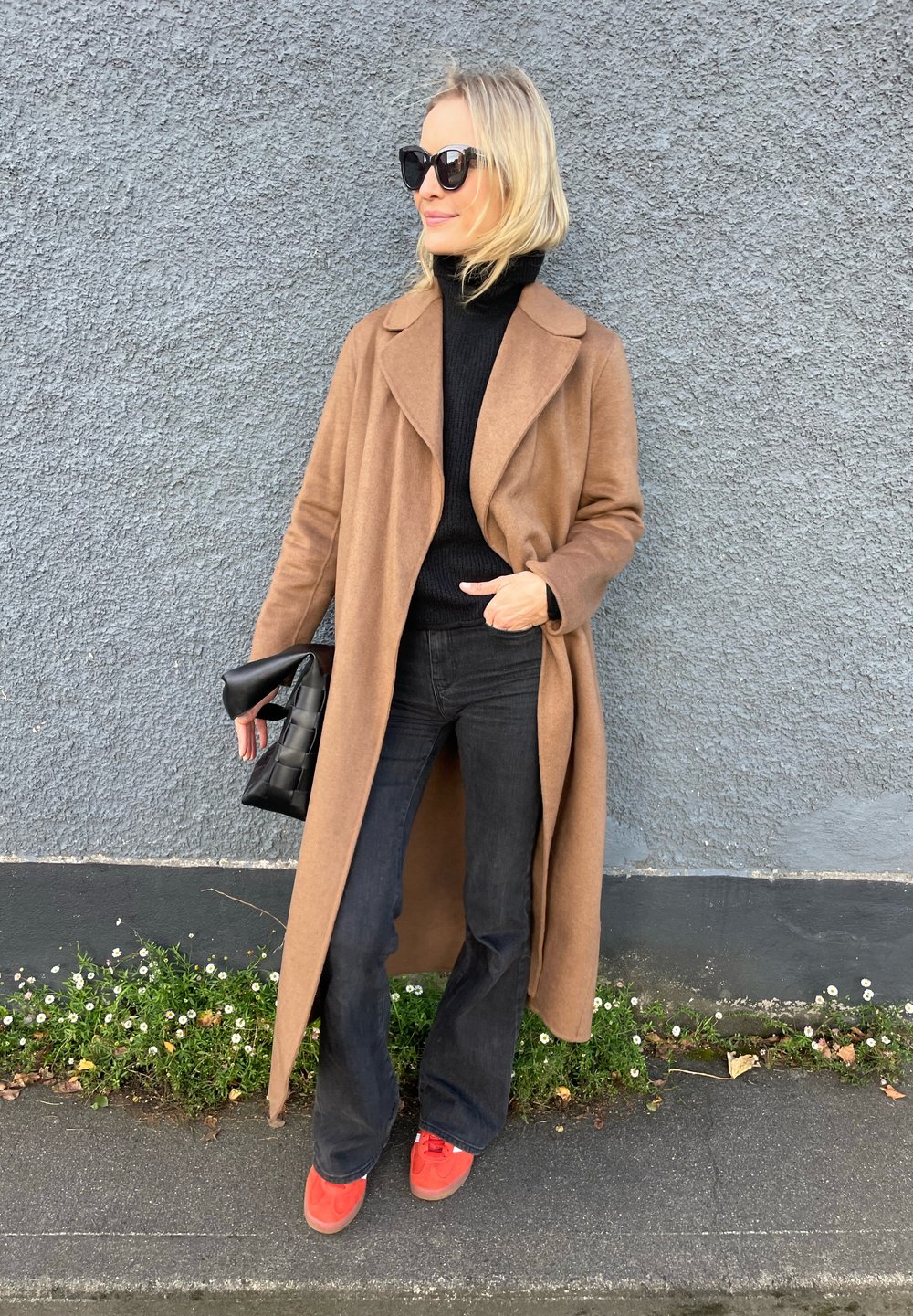 Brauner Wollmantel mit breitem Kragen, schwarzer Rollkragenpullover, weit geschnittene schwarze Jeans und rote Sneakers. Hält eine schwarze Handtasche. Graue Wand im Hintergrund.