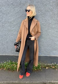 Brauner Wollmantel mit breitem Kragen, schwarzer Rollkragenpullover, weit geschnittene schwarze Jeans und rote Sneakers. Hält eine schwarze Handtasche. Graue Wand im Hintergrund.