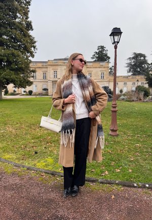 Mujer con abrigo beige, suéter blanco, bufanda estampada, pantalones negros, gafas de sol y sosteniendo un bolso blanco, de pie en un sendero en un jardín.