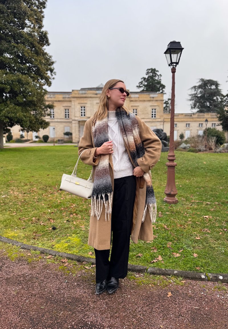 Femme portant un manteau beige, un pull blanc, une écharpe à motifs, un pantalon noir, des lunettes de soleil, et tenant un sac à main blanc, debout sur un chemin dans un jardin.