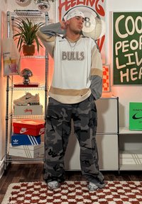 Biała i beżowa koszulka hokejowa z napisem "BULLS", szare spodnie dresowe w kamuflażu i szare sneakersy. Dodatki obejmują łańcuch i bandanę.