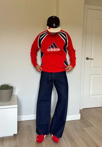Rød Adidas langermet skjorte med marineblå detaljer, kombinert med vide mørk denimbukser og røde sko. En enkel svart og hvit cap fullførte antrekket.