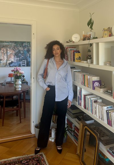 Camisa de manga larga azul claro, de corte holgado, combinada con pantalones negros de cintura alta. Zapatos negros de punta, bolso marrón de hombro, ambiente interior con libros.