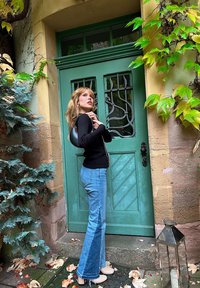 Maglione nero, jeans chiari a palazzo azzurri, scarpe crema. Porta verde con lavorazione metallica decorativa e muro di pietra, circondata da fogliame e lanterna.