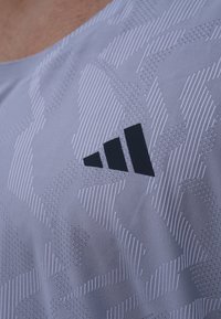 Grå treningsskjorte laget av teksturert stoff, med et subtilt geometrisk mønster. Svart logo tydelig vist på venstre side.