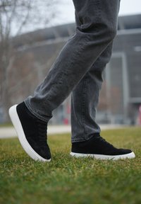 Baskets en daim noir avec semelles en caoutchouc blanc, finition texturée et lacets plats, portées avec un jean gris, debout sur l'herbe.