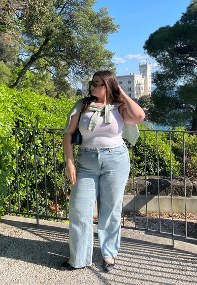 Una persona vestida con una camiseta blanca sin mangas y pantalones vaqueros de pierna ancha de un azul claro está de pie al aire libre, con un suéter de punto verde claro sobre los hombros, junto a la vegetación.
