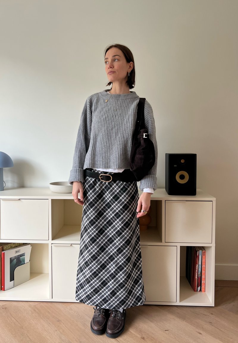 Pull gris côtelé, jupe longue à carreaux noirs avec accents blancs, ceinture noire, chaussures marron à lacets, portant un sac à bandoulière foncé.