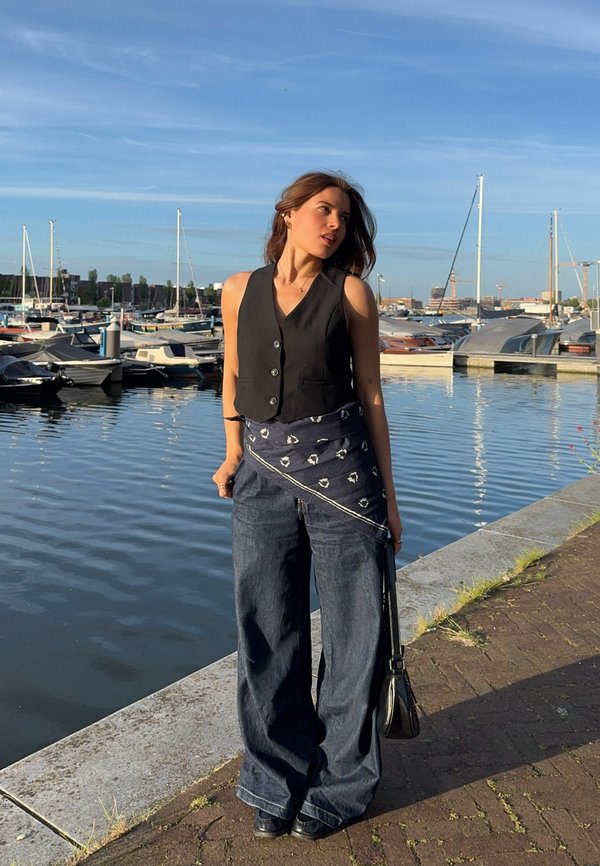 Femme debout près d'une marina avec des bateaux, portant un gilet noir, un jean à jambes larges et un cache-écharpe à motifs, regardant de côté sous la lumière du soleil.