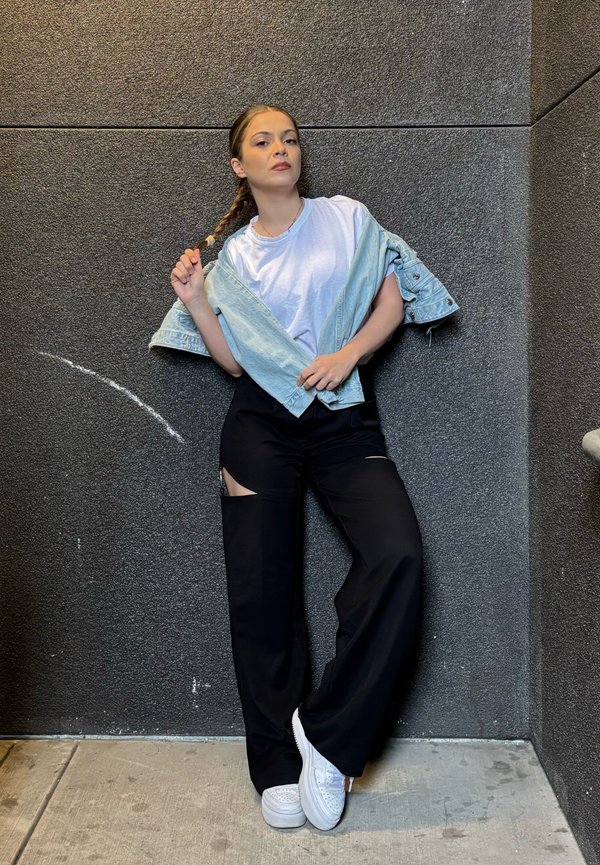 Casaco de ganga azul claro, t-shirt branca e calças largas pretas com recortes. Modelo está encostado a uma parede escura texturizada, usando sapatilhas brancas.