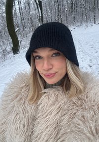 Bonnet noir en tricot, manteau beige clair duveteux avec de longues fibres, col montant, et cheveux blonds mi-longs sur fond de forêt enneigée.