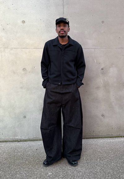 Hombre de pie contra una pared de cemento, vestido con gorra negra con broche, chaqueta negra, pantalones anchos y oscuros, y zapatos negros, con las manos en los bolsillos.