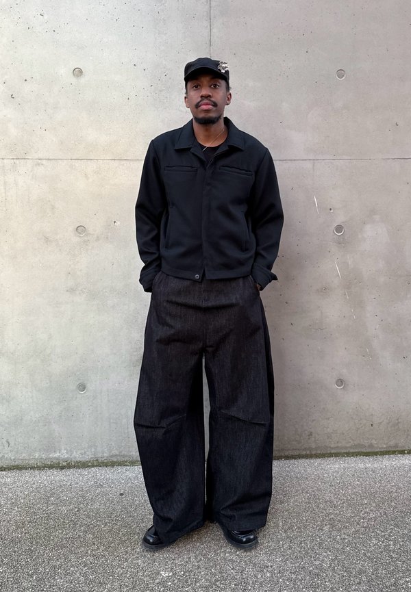 Άνδρας στέκεται δίπλα σε τσιμεντένιο τοίχο φορώντας μαύρο καπέλο με καρφίτσα, μαύρο σακάκι, φαρδιά σκουρόχρωμα παντελόνια και μαύρα παπούτσια, με τα χέρια στις τσέπες.