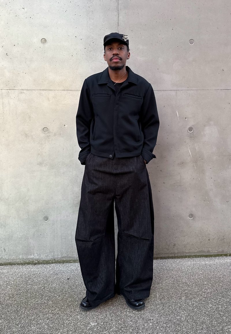 Homme debout contre un mur en béton portant une casquette noire avec une broche, une veste noire, un pantalon large et sombre, et des chaussures noires, les mains dans les poches.