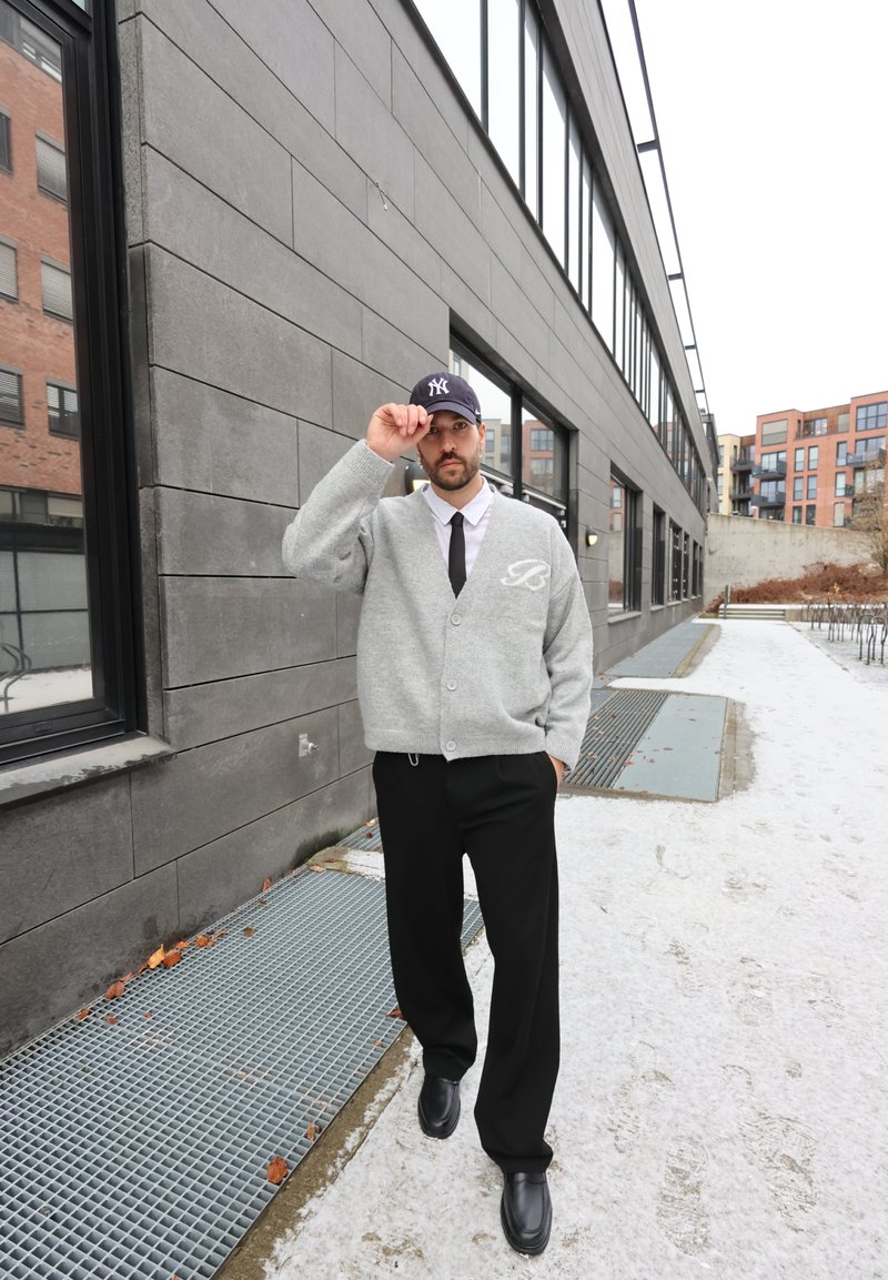 Cardigan gris avec une lettre "B", chemise blanche, cravate noire et pantalon noir. Casquette noire avec logo blanc ; chaussures noires brillantes. Fond urbain.