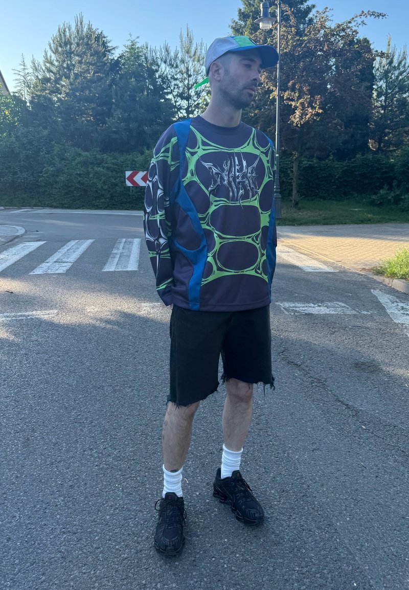 Homme portant une chemise à manches longues à motif, un short noir, des chaussettes blanches, des chaussures noires et une casquette bicolore, debout dans une rue près d'un passage pour piétons.