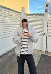 Moletom cinza com fecho e capuz com cordão, usado sobre uma t-shirt às riscas brancas e pretas. Calças pretas e óculos de sol completam o look.
