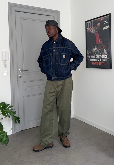 Hombre que lleva una chaqueta vaquera oscura, pantalones verde oliva, botas marrones y una gorra hacia atrás, de pie cerca de una puerta gris cerrada en una habitación blanca.