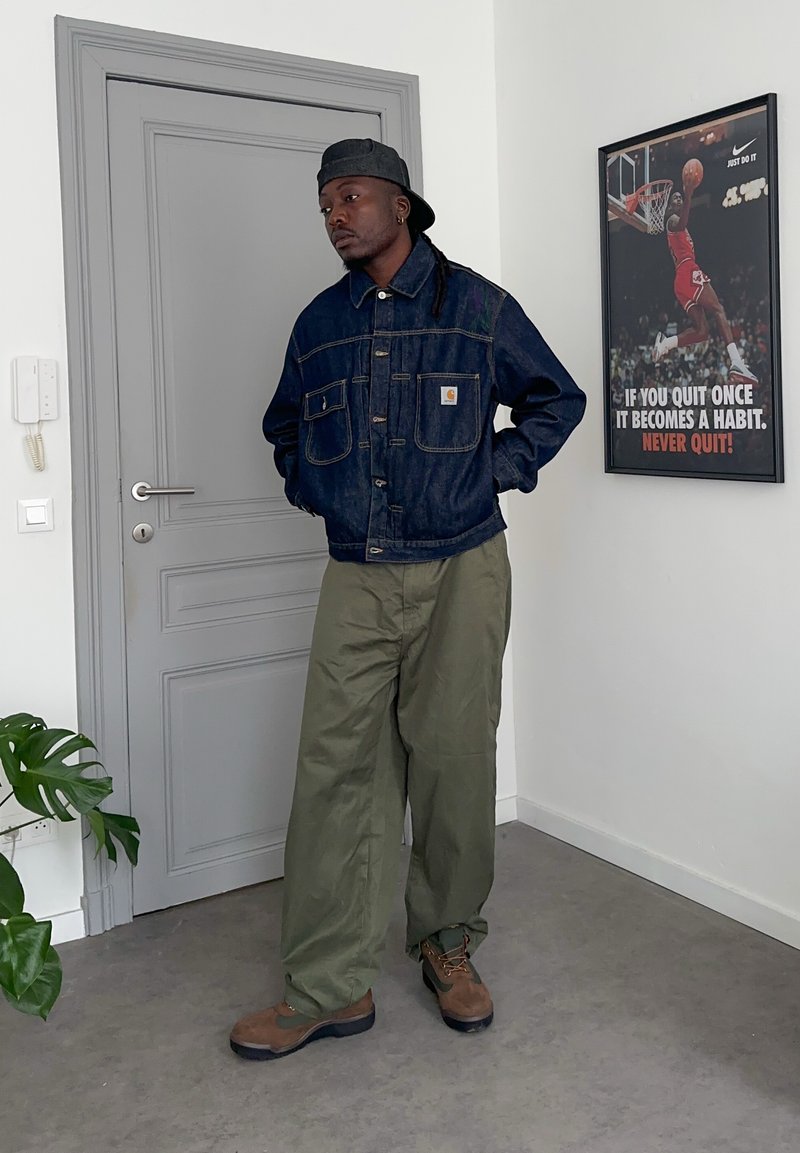 Ein Mann trägt eine dunkle Jeansjacke, olivgrüne Hose, braune Stiefel und eine rückwärts getragene Kappe und steht nahe einer geschlossenen grauen Tür in einem weißen Raum.