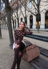 Femme en haut marron à pois et collants assise sur un banc de parc à côté d'un grand sac à main marron, avec des arbres sans feuilles et un bâtiment historique en arrière-plan.