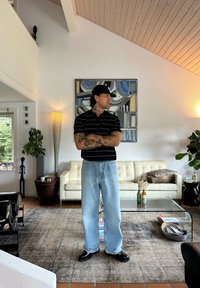 Hombre con tatuajes y una gorra negra está de pie con los brazos cruzados en una sala de estar moderna con un sof�á blanco, mesa de vidrio y arte abstracto en la pared.