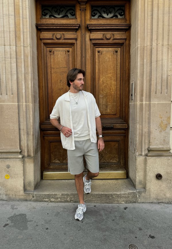 Camisa de manga corta de color claro sobre una camiseta blanca, combinada con pantalones cortos a rayas y zapatillas grises, con fondo de puertas de madera ornamentadas.