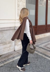 Brun tørklæde hængende over en hvid sweater, sorte bukser og brune sneakers. Holder en tekstureret, olivengrøn håndtaske. Indendørs arkitektonisk baggrund.