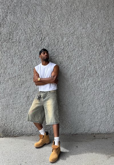 Camisa sin mangas blanca, pantalones cortos holgados verde claro, botas de trabajo marrones y calcetines blancos. Diadema negra y llaves visibles en un gancho del cinturón.