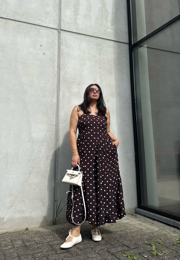 Femme portant une robe à pois noir et blanc, des chaussures blanches, des lunettes de soleil, et tenant un petit sac à main blanc, debout près d'un mur en béton gris et d'une fenêtre en verre.