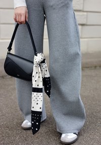 Personne portant un pantalon large gris et des baskets blanches, tenant un sac à main noir avec un foulard noir à pois blancs attaché à la poignée.