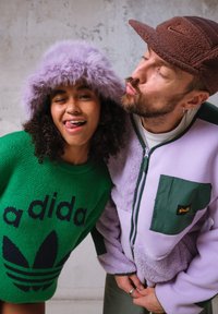 Pulôver verde com logotipo azul-escuro, gorro roxo felpudo e puffer leve roxo com detalhes em verde nos bolsos. Cores vibrantes e texturizadas.