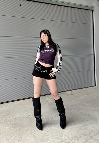 Joven vestida con shorts negros, una blusa larga y transparente debajo de una camiseta púrpura, cinturón con tachuelas y botas negras hasta la rodilla posa frente a una pared gris.