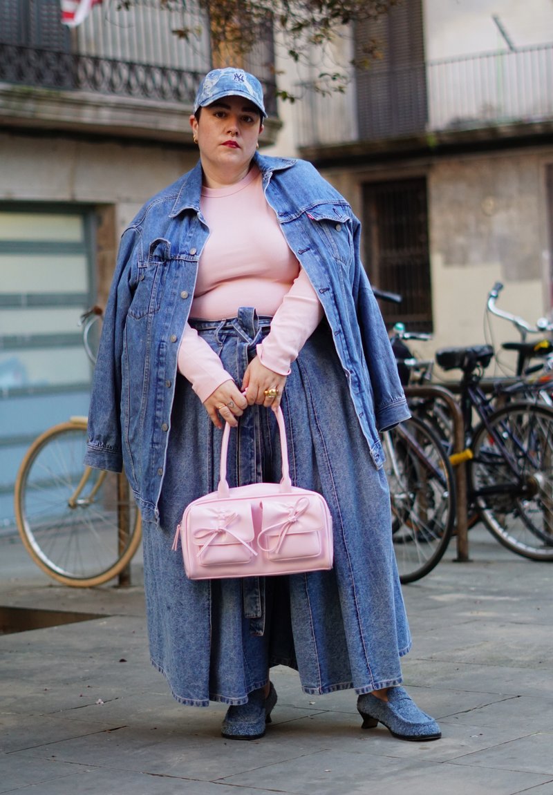 Femme portant une veste en denim, une longue jupe en denim, un haut rose et une casquette bleue, tenant un sac à main rose, debout dans une rue avec des vélos en arrière-plan.