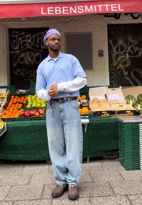 Chemise à rayures bleues à manches courtes avec des poignets blancs, jean baggy bleu clair et sabots marron. Produits frais exposés dans des caisses vertes derrière.
