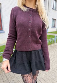 Femme portant un cardigan violet boutonné, une jupe noire en plusieurs couches et des collants à motifs de dentelle, debout sur un chemin pavé près de bâtiments blancs.