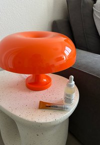 Πορτοκαλί πλαστικό φωτιστικό μανιταριού πάνω σε υφασμάτινο λευκό τραπέζι. Κοντά υπάρχουν ένα διαφανές μπουκάλι σταγόνας και μια ασημένια σωλήνα κίνησης.