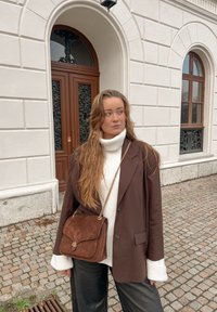 Sac à bandoulière en daim marron avec un rabat, une quincaillerie dorée et une bandoulière chaîne. Porté avec un blazer marron surdimensionné et un pull à col roulé blanc.