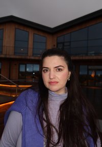 Lila Strickpullover über einem hellgrauen Rollkragen, langes dunkles Haar und dezenter Schmuck, vor einem modernen Holzgebäude.