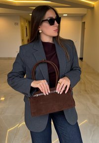 Sac à main en daim marron avec des poignées arrondies, une fermeture éclair argentée en accent et une forme structurée. Porté avec un blazer gris et un pantalon foncé.