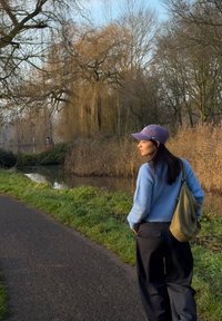 Pull bleu clair en tricot, pantalon noir ample et casquette violette. Chemin d'herbe verte à côté d'un cours d'eau avec des hautes herbes et des arbres.