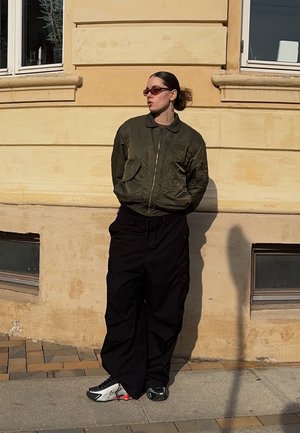 Chaqueta bomber de color verde oliva, pantalones cargo negros y zapatillas en blanco y negro. Persona de pie contra una pared beige con ventana.