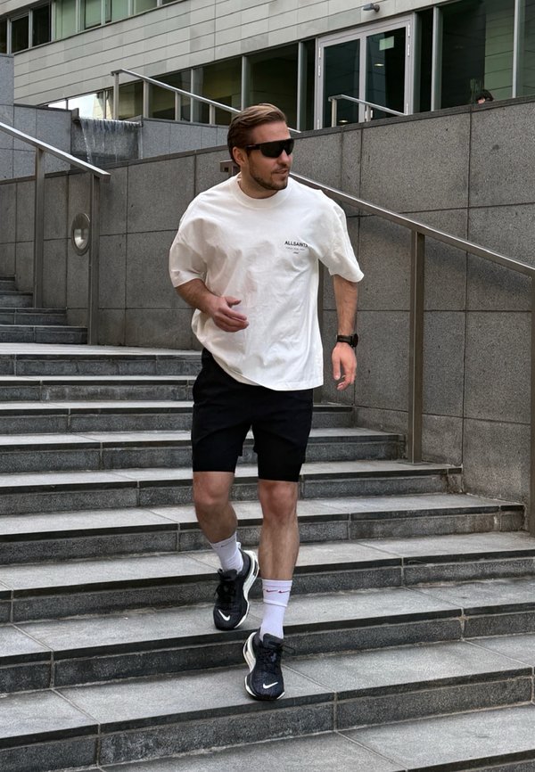Bărbat purtând ochelari de soare, tricou alb, pantaloni scurți negri și adidași, coborând o scară exterioară lângă un zid de piatră gri cu balustradă.
