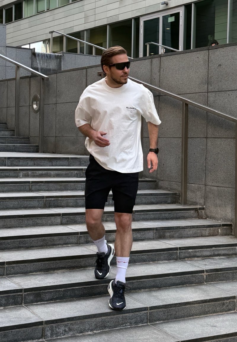 Homme portant des lunettes de soleil, un t-shirt blanc, un short noir et des baskets qui descend des escaliers extérieurs à côté d'un mur en pierre grise avec des rampes.