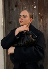 Een zwarte, textuur afgewerkte handtas wordt dicht tegen een donkere jas gehouden. De achtergrond toont een rustieke houtenwand met zichtbare knopen en slijtage.